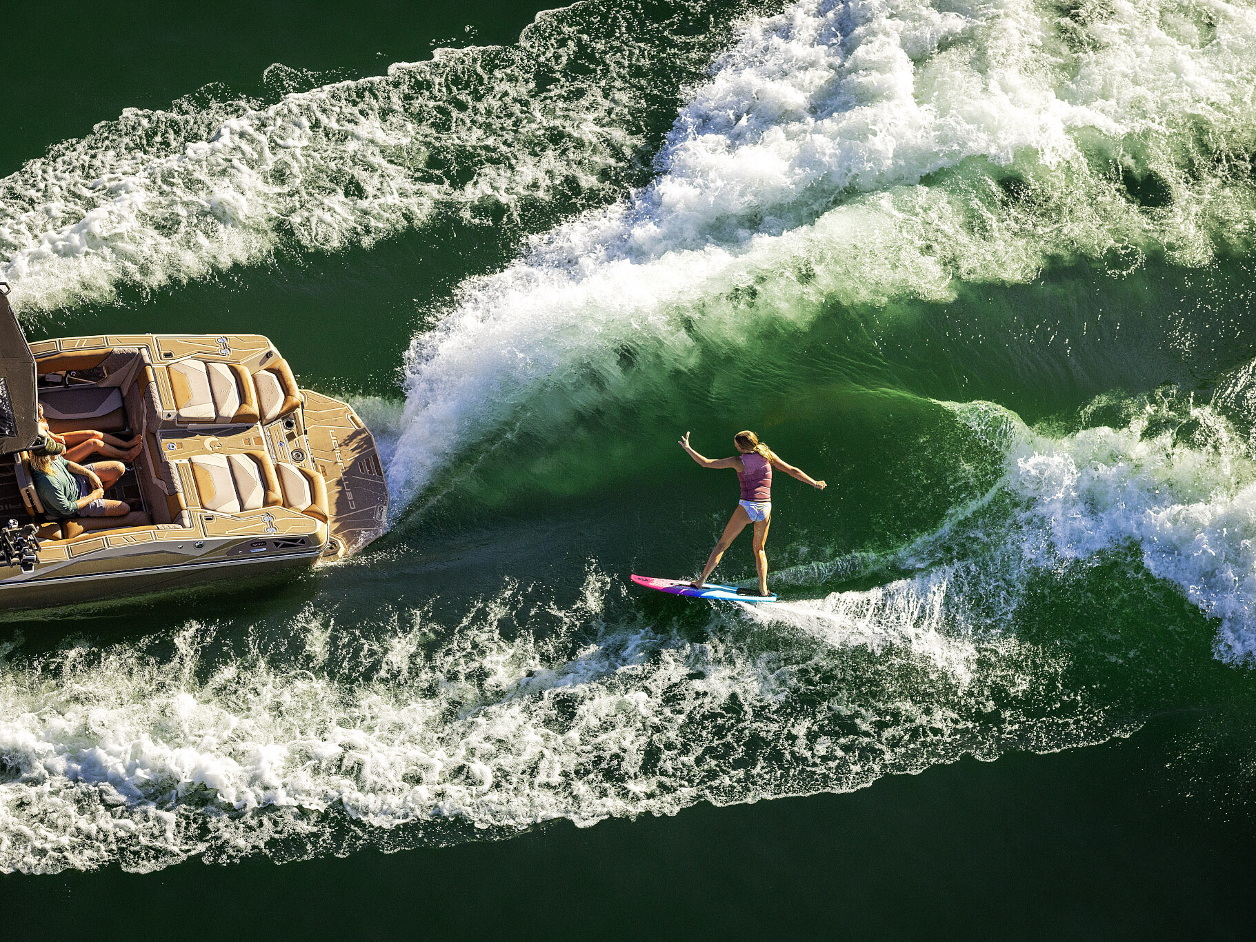 A person in swimwear wake surfs behind a Centurion Ri Series motorboat on green water, skillfully riding the waves created by the boat.