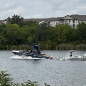 A person wakeboards behind a motorboat on a river, with trees and residential buildings visible in the background under a cloudy sky—capturing an action-packed moment reminiscent of the energy at Centurion Cowtown Wake Fest.