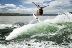 A person in a black wetsuit rides a surfboard, jumping off a green wave near a California Boat Company vessel under a partly cloudy sky.
