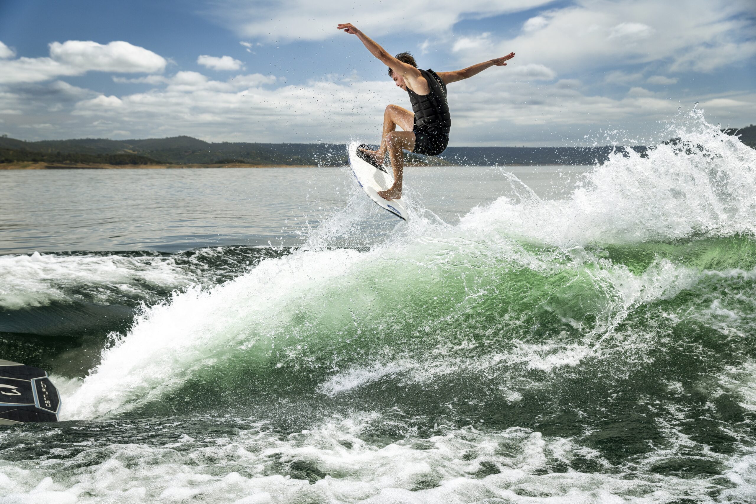 A person in a black wetsuit rides a surfboard, jumping off a green wave near a California Boat Company vessel under a partly cloudy sky.