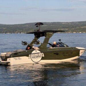 Green and white speedboat with “World Wake Surfing” logo floats on a lake; two people are seated inside, hills and water visible in the background, capturing the excitement of the upcoming Centurion Surf Classic presented by Canandaigua Boatworks.
