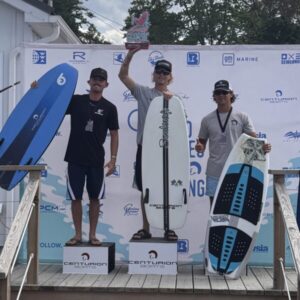 Three men stand on a winners’ podium holding surfboards at the Centurion Surf Classic Surf Finals; the center man holds a trophy. Event sponsor logos, including Canandaigua Boatworks, are visible on the background banner.
