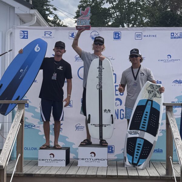 Three men stand on a winners&rsquo; podium holding surfboards at the Centurion Surf Classic Surf Finals; the center man holds a trophy. Event sponsor logos, including Canandaigua Boatworks, are visible on the background banner.