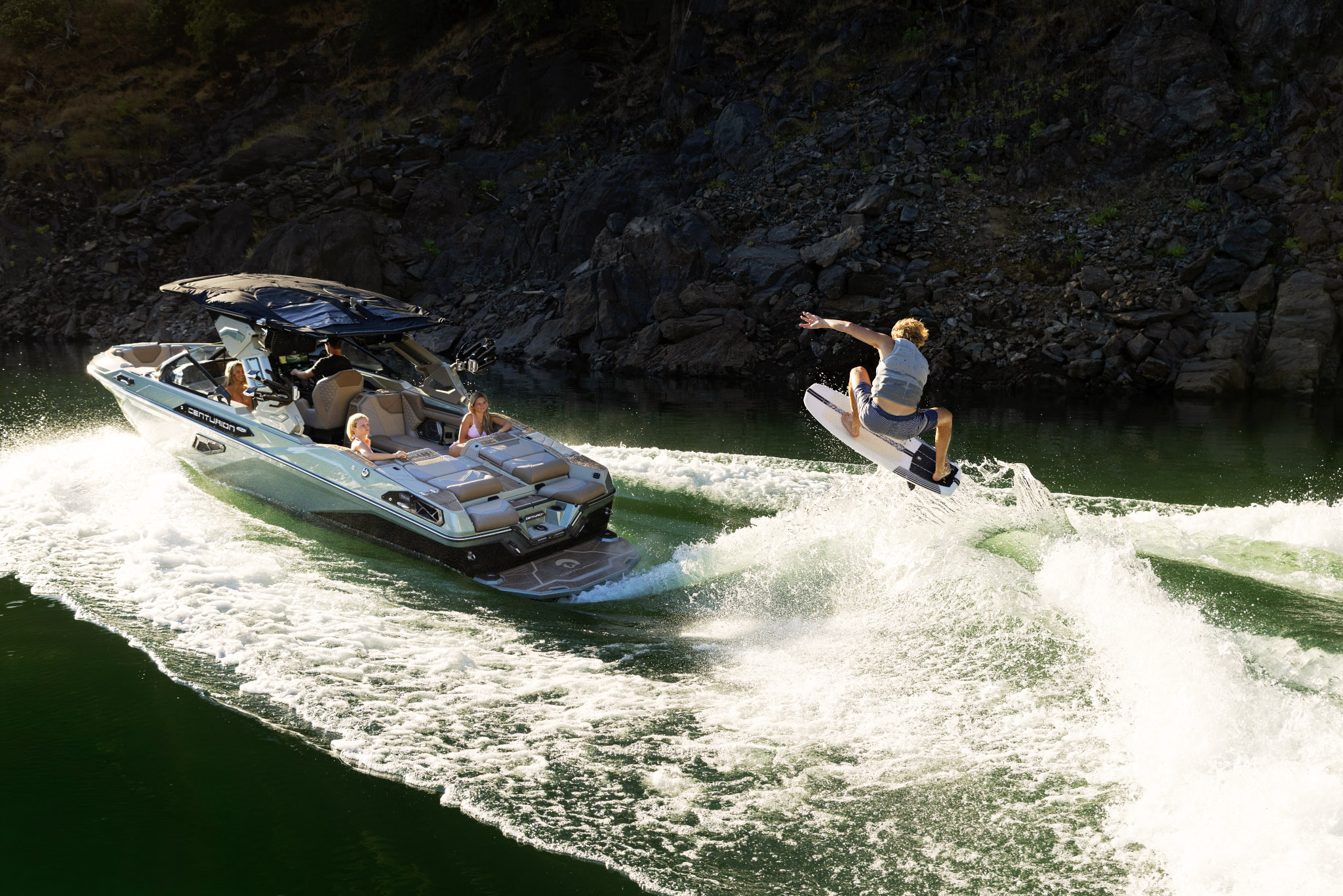 A man rides a wakeboard and jumps over the water behind a GM Marine-powered motorboat carrying three people on a lake surrounded by rocky terrain, setting the stage for an exhilarating scene worthy of the 2026 World Wake Surfing Championship.