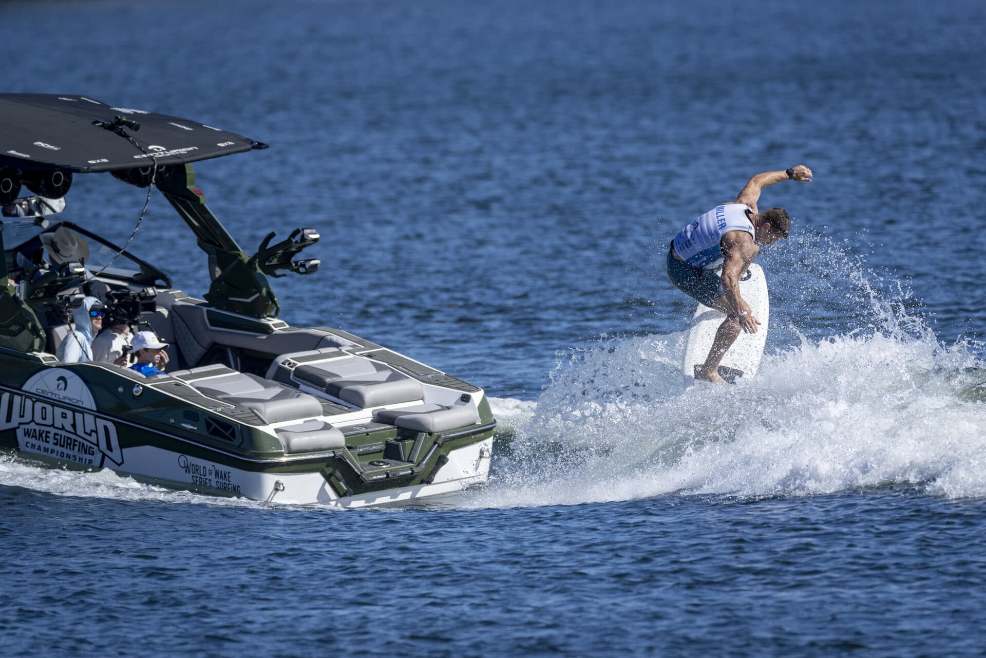 A person wake surfing behind a boat on a calm blue lake, as two watch from inside during Day 1 Recap of the Centurion World Wake Surfing Championship.