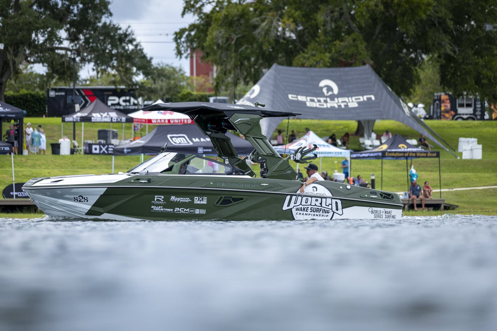 A green and white wakeboarding boat with “World Wake Surfing” branding floats on the water near event tents at the Wake Surfing Championship on a grassy shore.